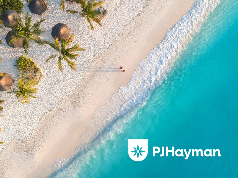 Aerial beach holiday photograph of clear blue water, waves and soft sandy beach with palm trees and covers with people walking across the sand, with the PJHayman logo