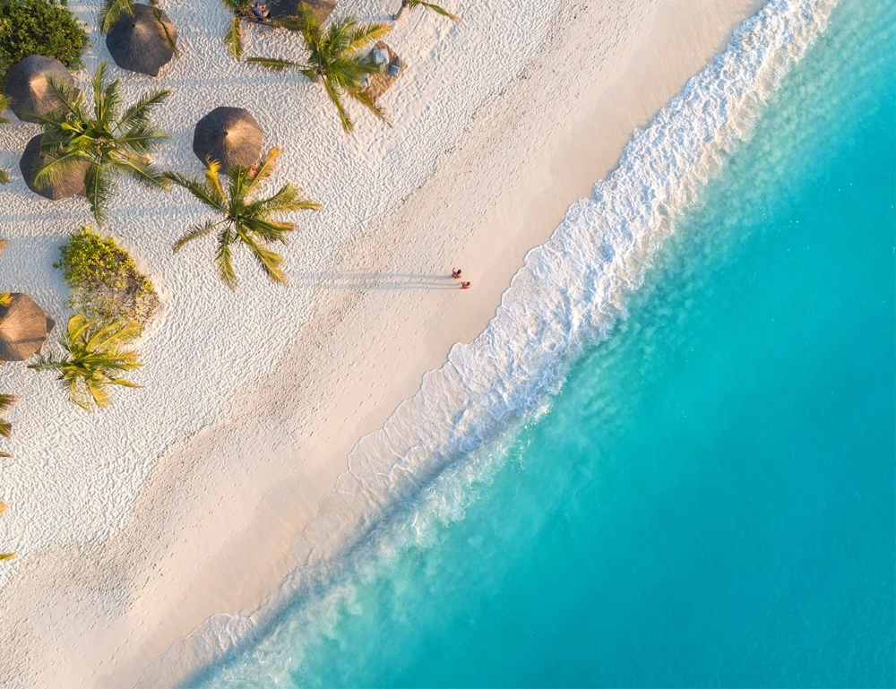 Aerial beach holiday photograph of clear blue water, waves and soft sandy beach with palm trees and covers with people walking across the sand