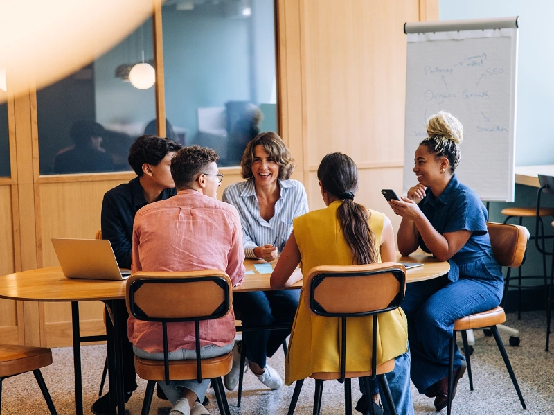 Group of young adults in open working space around a table talking and laughing using phones and laptop with whiteboard
